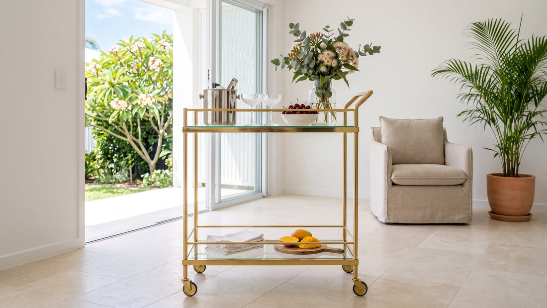 A gold and glass bar cart styled with eucalyptus stems, a bowl of cherries and an ice bucket in a living room open to a sunlit Noosa garden