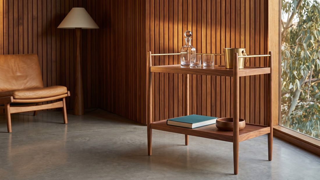 A walnut mid-century bar cart with tapered legs and a brass rail against a timber batten wall