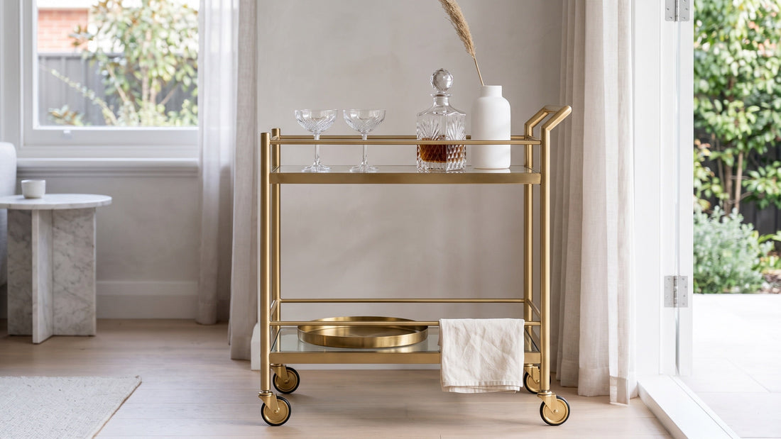 A brushed gold and glass two-tier bar cart catching afternoon window light in an Adelaide villa living room
