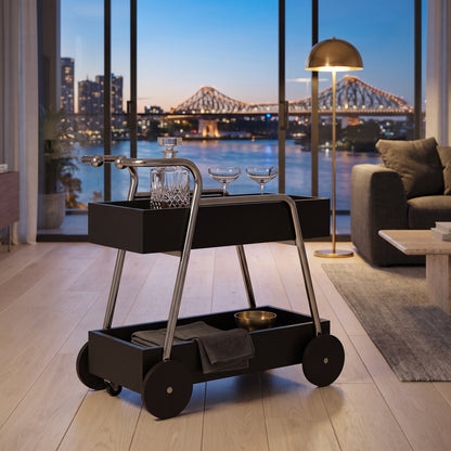 The Carlton Mid-Century Solid Wood Drinks Trolley in black beside a sofa in a Brisbane riverside apartment at dusk, the Story Bridge through the glass