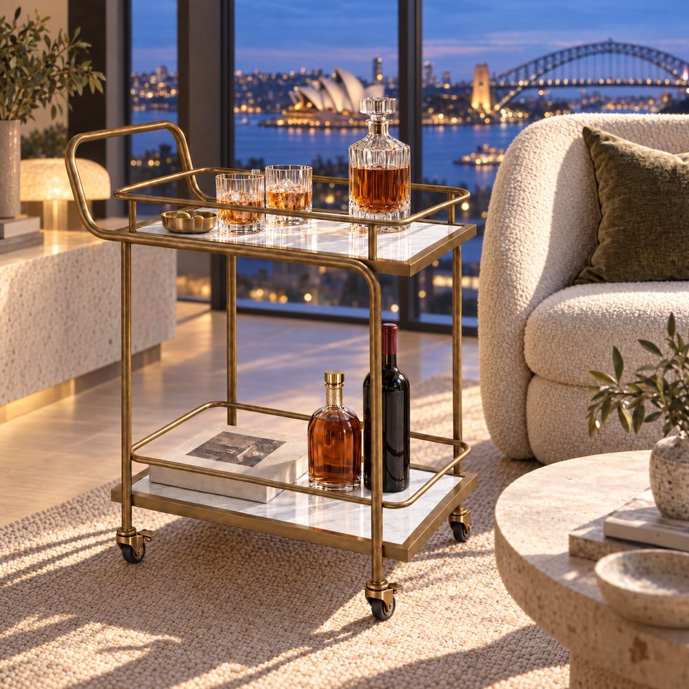 The Subiaco Nordic Service Trolley in Aged Brass with white marble-look shelves in a Sydney apartment at dusk, the Opera House and Harbour Bridge through the window