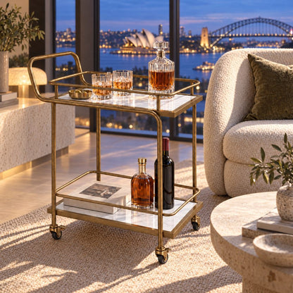The Subiaco Nordic Service Trolley in Aged Brass with white marble-look shelves in a Sydney apartment at dusk, the Opera House and Harbour Bridge through the window
