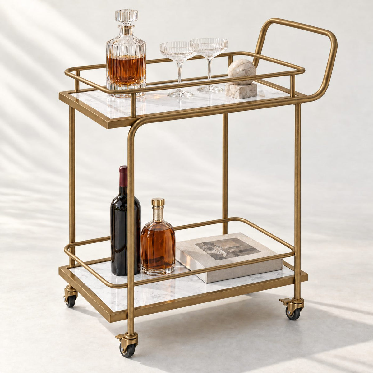 The Subiaco Nordic Service Trolley in Aged Brass with white marble-look board shelves, styled with a decanter and coupe glasses, on a white background