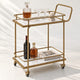 The Subiaco Nordic Service Trolley in Aged Brass with white marble-look board shelves, styled with a decanter and coupe glasses, on a white background